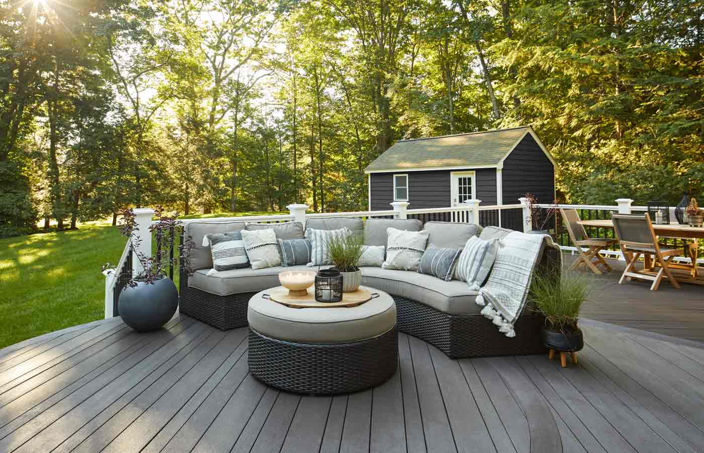Modern outdoor deck with a curved sectional sofa, round ottoman, potted plants, and a dining table set, surrounded by trees and a small shed in the background.